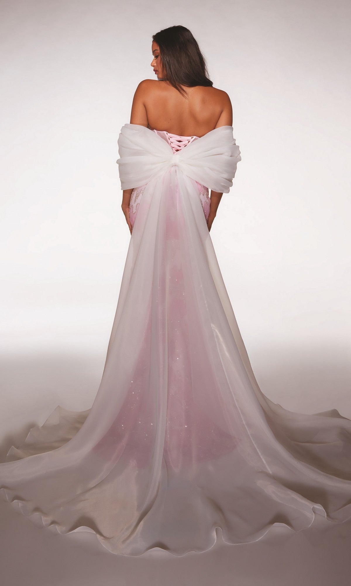A woman faces away from the camera in the Alyce 62187 long prom dress, featuring a strapless pink gown with a sheer corset bodice, a large removable white bow, and a long flowing train against a plain background.